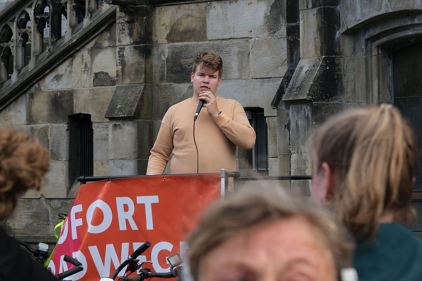 Ben Jansen auf einer Demonstration Ben Jansen spricht bei der Mahnwache vor dem Aachener Rathaus in ein Mikrofon. Vor ihm hängt ein orange-rotes Banner des Radentscheids.