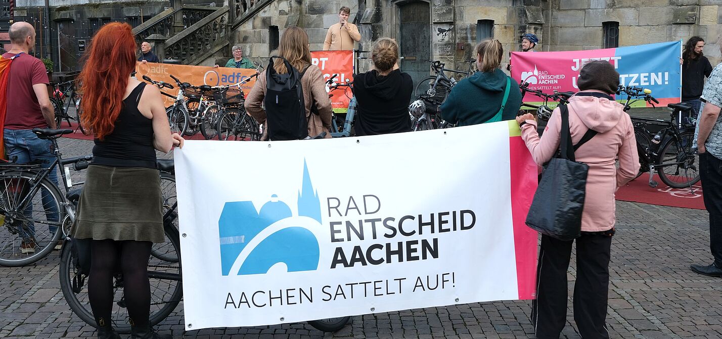 Teilnehmende einer Mahnwache vor dem Aachener Rathaus stehen mit Fahrrädern und Bannern von Radentscheid Aachen und ADFC vor einem Redner mit Mikrofon.