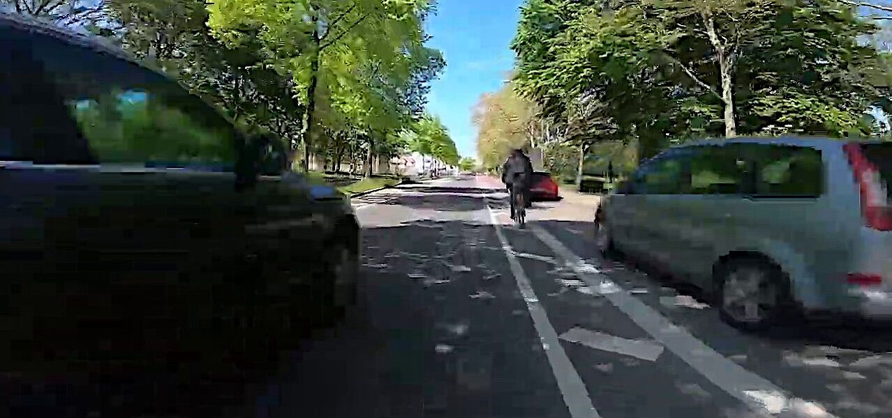 Ein Fahrradfahrer fährt neben einem zugeparkten Radstreifen, während sich ein weiteres Kfz von hinten nähert