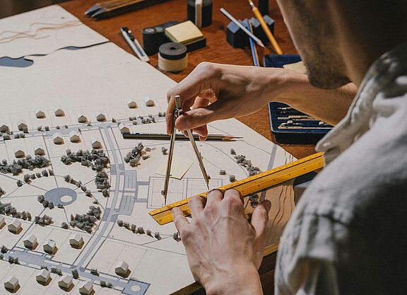 Ein Stadtplaner bei der Arbeit Ein Mann im Hemd ist von hinten zu sehen und zeichnet mit Lineal und Zirkel etwas auf einen Stadtplan.