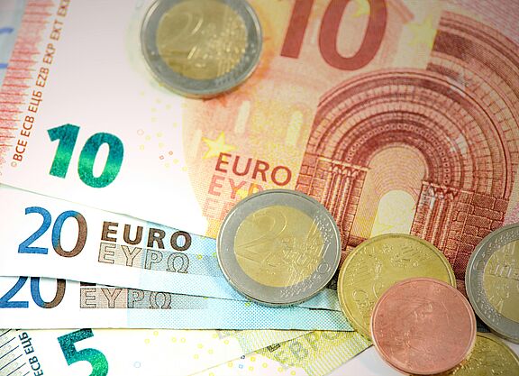 Euro-Bargeld in Scheinen und Münzen Es sind mehrere Geldscheine zu sehen. Ein Zehn-Euro-Schein liegt auf mehreren Zwanzig-Euro- und einem Fünf-Euro-Schein. Außerdem liegen einige Münzen auf den Scheinen verstreut.