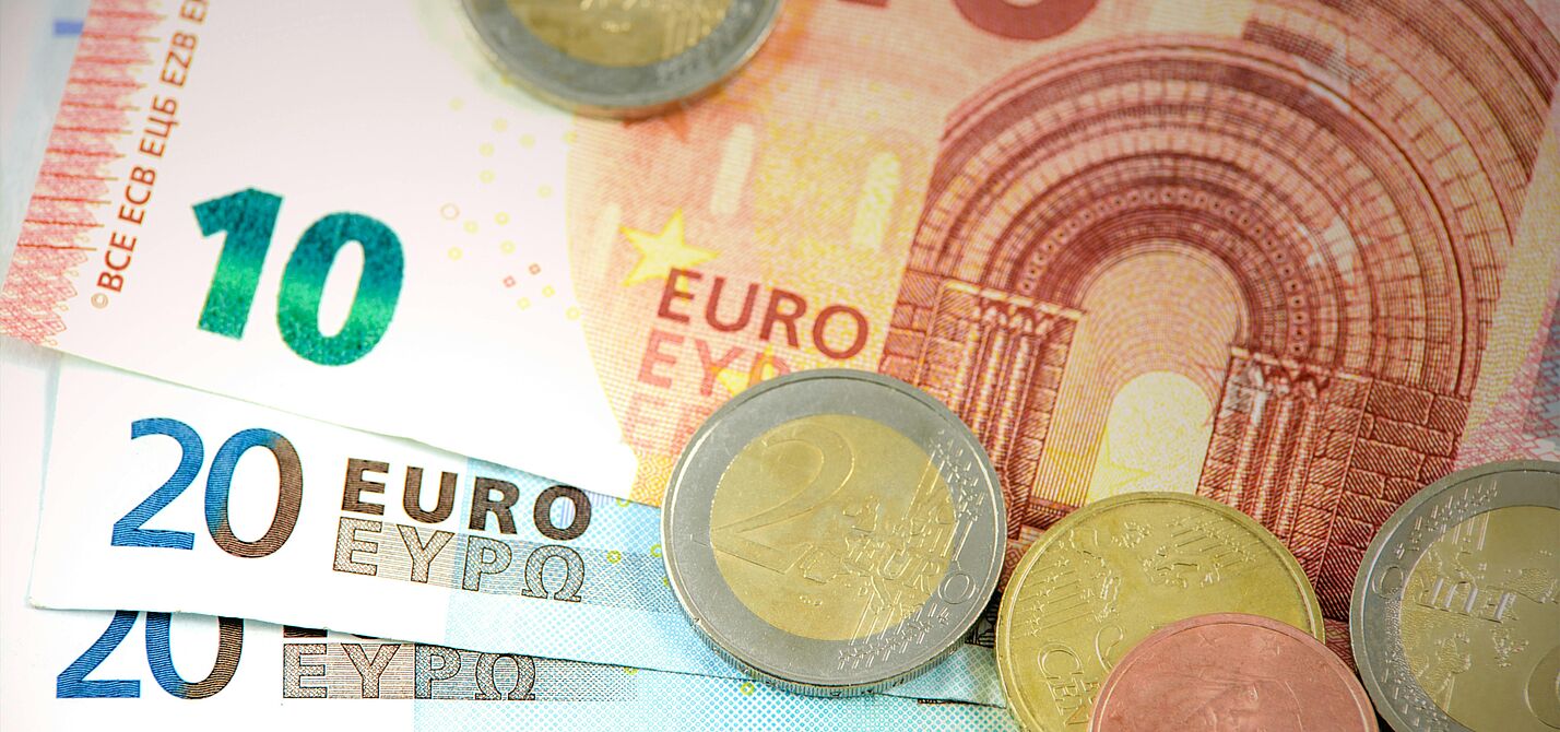Euro-Bargeld in Scheinen und Münzen Es sind mehrere Geldscheine zu sehen. Ein Zehn-Euro-Schein liegt auf mehreren Zwanzig-Euro- und einem Fünf-Euro-Schein. Außerdem liegen einige Münzen auf den Scheinen verstreut.