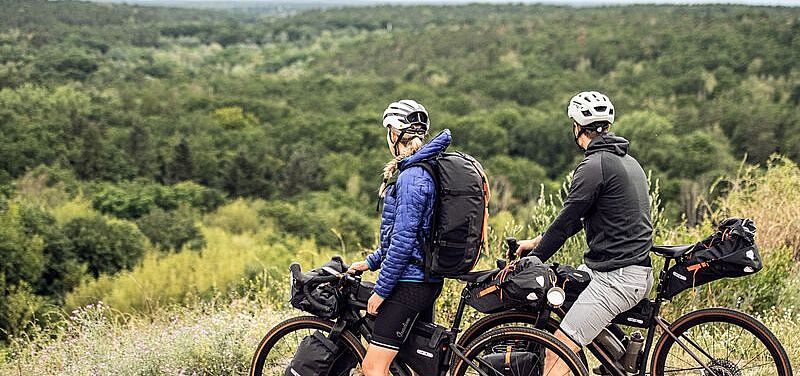 Eine Radfahrerin und ein Radfahrer stehen auf einer Anhöhe un blicken auf einen Wald. 