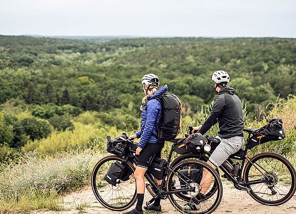 Bikepacking Eine Radfahrerin und ein Radfahrer stehen auf einer Anhöhe un blicken auf einen Wald.