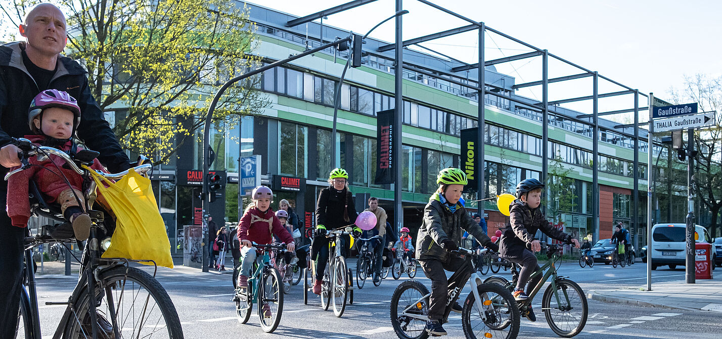 Eine Gruppe von Kindern fährt auf ihrer Rädern auf der Straße. Sie werden von Erwachsenen begleitet.