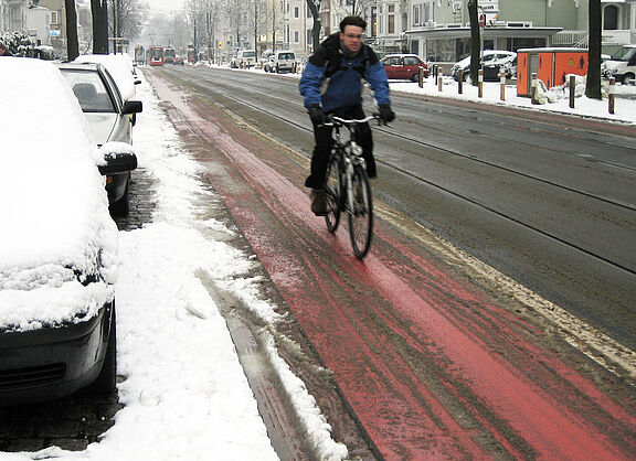 Radfahren im Winter Ein Radfahrer in Winterkleidung fährt auf einem rot markierten Radfahrstreifen neben einer Straße, der weitgehend von Schnee befreit ist, während am Straßenrand und auf geparkten Autos Schnee liegt.